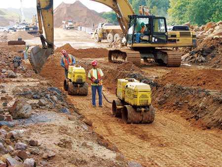 Joe Cox and Rita Tonkin compact backfill over a drainage pipe in East Beckley, as Hoe Operator Alfonso Torres places a new layer of backfill material behind them. (Photo by Carl Thiemann)